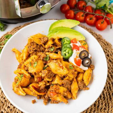 A dinner bowl with cheesy taco pasta made in the instant pot garnished with avocado, sour cream and sliced jalapenos and black olives.