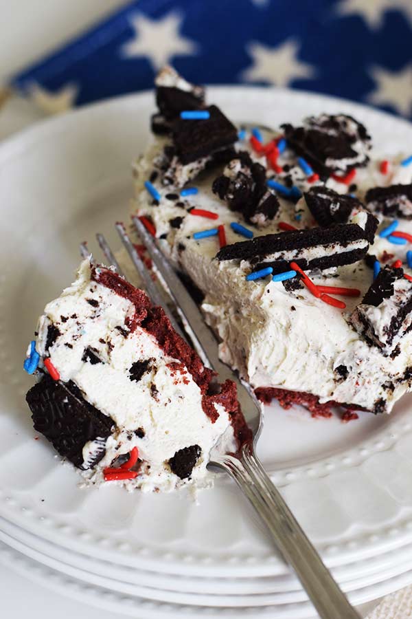 A slice of a no bake fireworks oreo cheesecake bar with red, white, and blue sprinkles and chopped oreos on top with a fork breaking off one bite