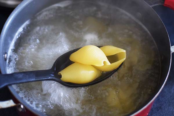 A closeup of large cooked pasta shells over a pot of boiling water in a large black serving spoon 