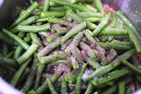 A close up of fresh and bright green green beans with salt and pepper in the instant pot with the broth, onions, and bacon