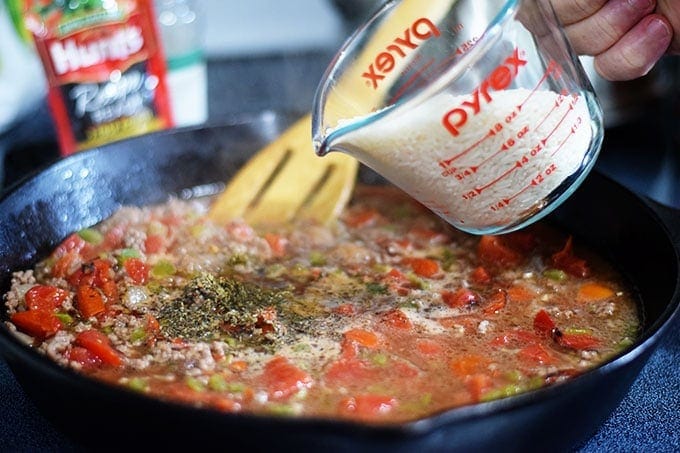 A closeup of the tomatoes, ground beef, onions, peppers, and seasonings cooking in a black cast iron skillet with a glass measuring cup filled with dry rice bring poured into the wet mixture