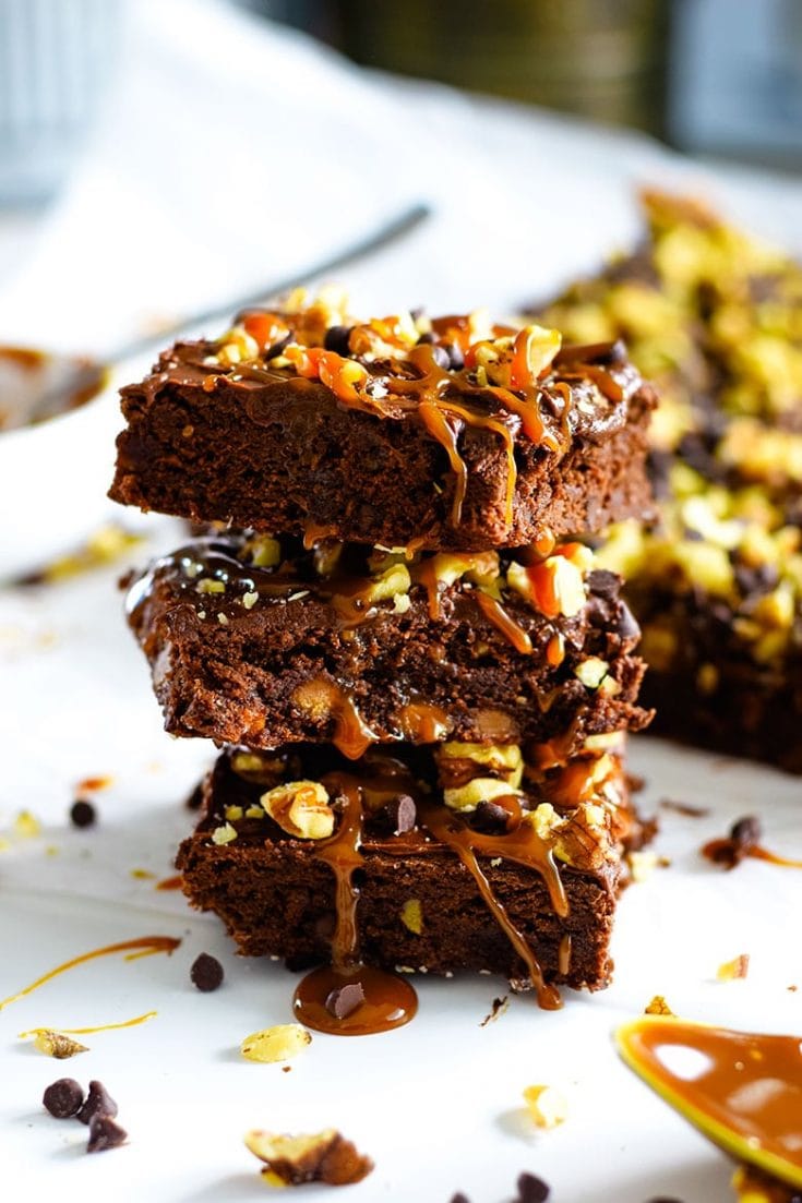 A stack of these frosted chocolate turtle brownies with caramel and chopped pecans