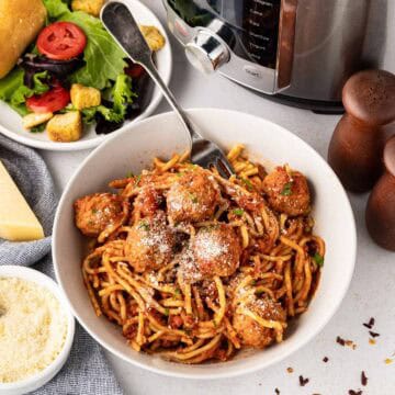 A bowl of spaghetti and meatballs, topped with grated Parmesan cheese and chopped parsley with a instant pot in the background.