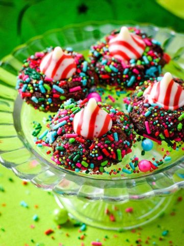 A close up of four chocolate candy cane Hershey's kiss cookies with Christmas sprinkles on a glass platter