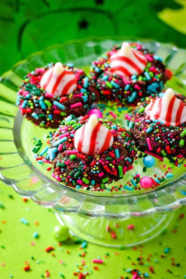 A close up of four chocolate candy cane Hershey\'s kiss cookies with Christmas sprinkles on a glass platter