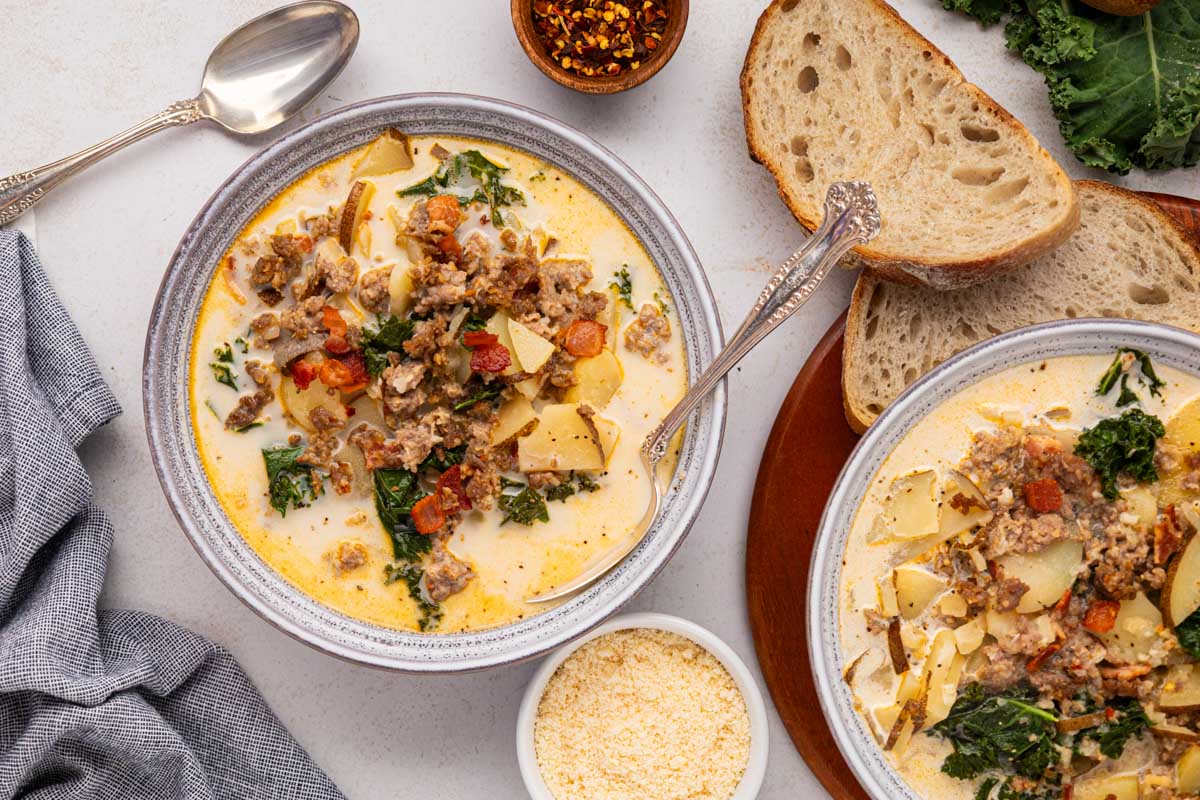 Two bowls of copycat olive garden Zuppa Toscana soup on a table with crusty bread, parmesan cheese, and crushed red pepper flakes for serving.