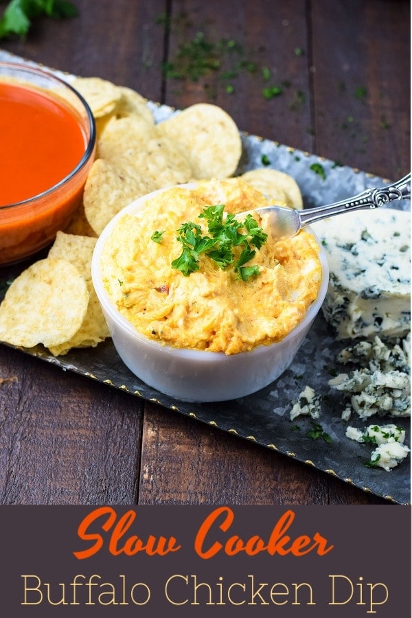 A tray with a bowl of buffalo sauce, tortilla chips, crumbled blue cheese, and a white bowl filled with slow cooker buffalo chicken dip to be served as a game day appetizer