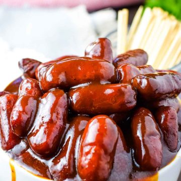 A white bowl filled with these crock pot barbecue little smokies smothered in bbq sauce with a cup of toothpicks for serving in the background