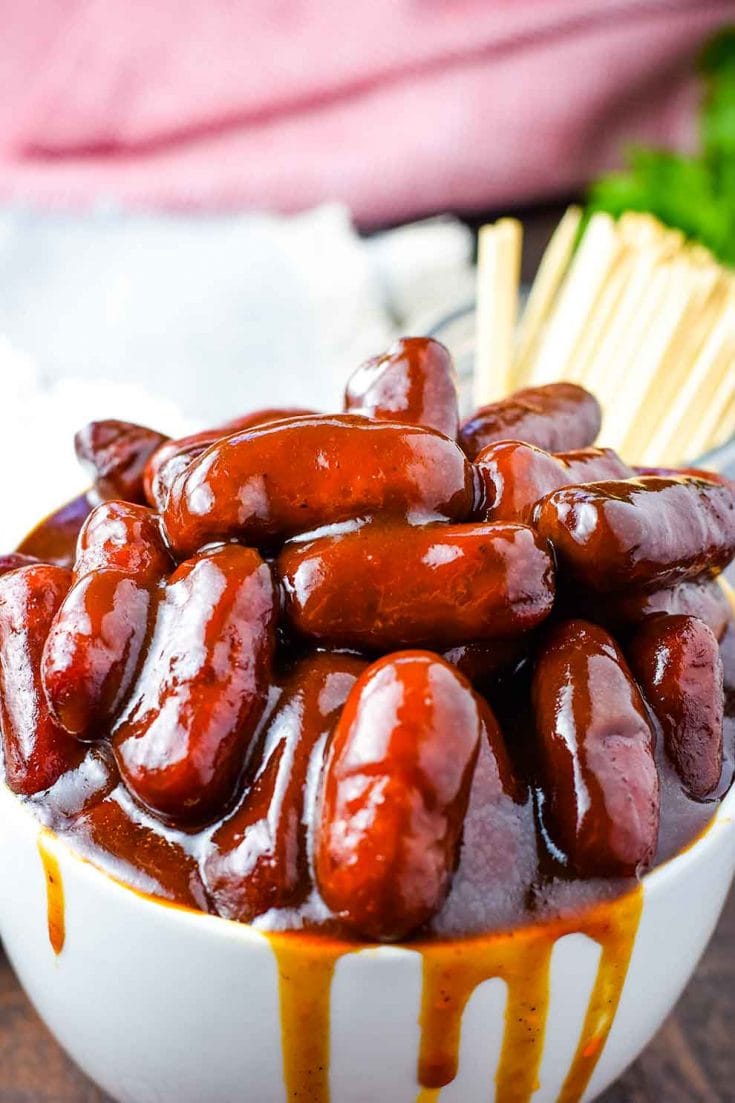 A white bowl filled with these crock pot barbecue little smokies smothered in bbq sauce with a cup of toothpicks for serving in the background