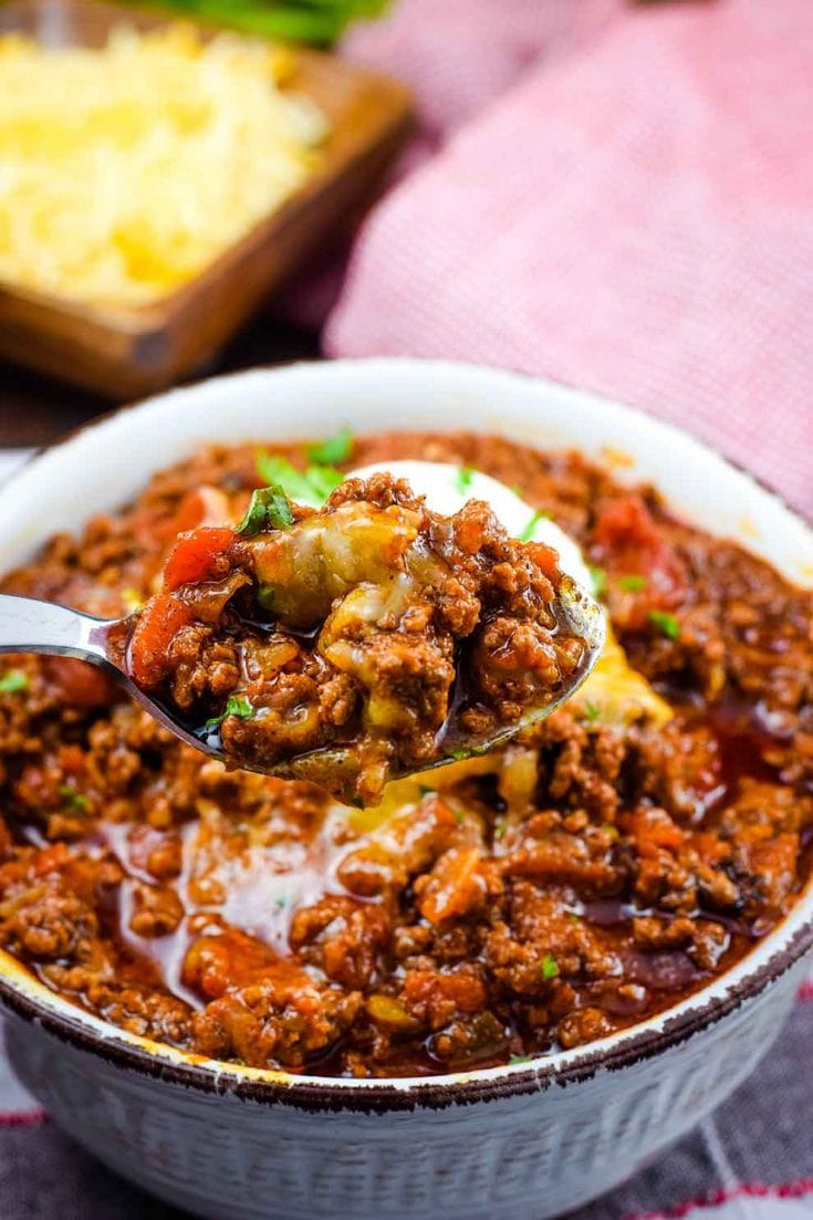 A bowl of this low-carb no-bean chili recipe topped with shredded cheese, sour cream, and parsley with a spoonful of this homemade chili in the forefront