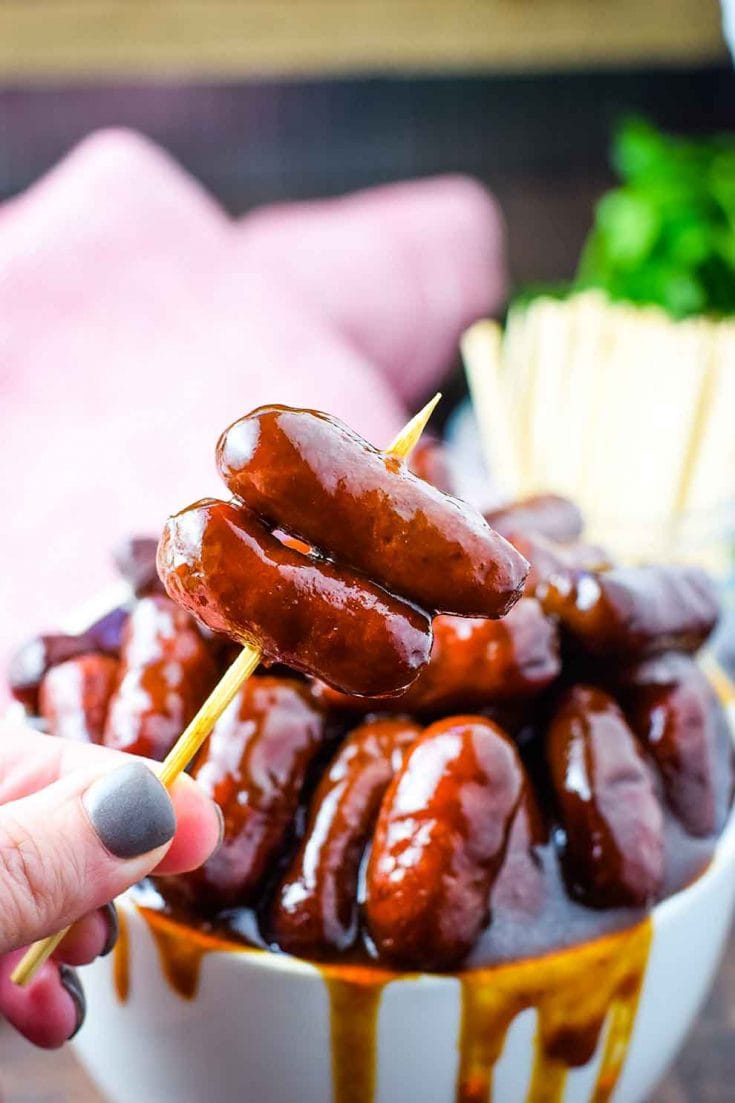 A white bowl filled with these crock pot barbecue little smokies smothered in bbq sauce with a cup of toothpicks for serving in the background