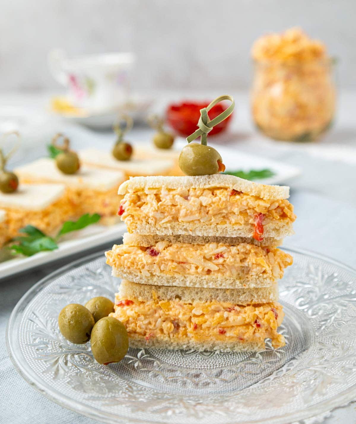 Pimento cheese sandwiches cut in triangles and stacked with an olive and wooden pic on top. 