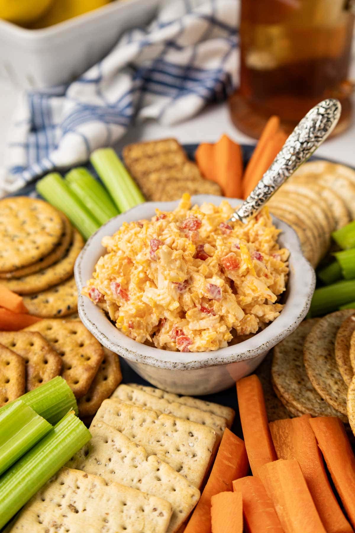 Pimento cheese in a bowl on a round wooden tray with crackers, celery and carrots.
