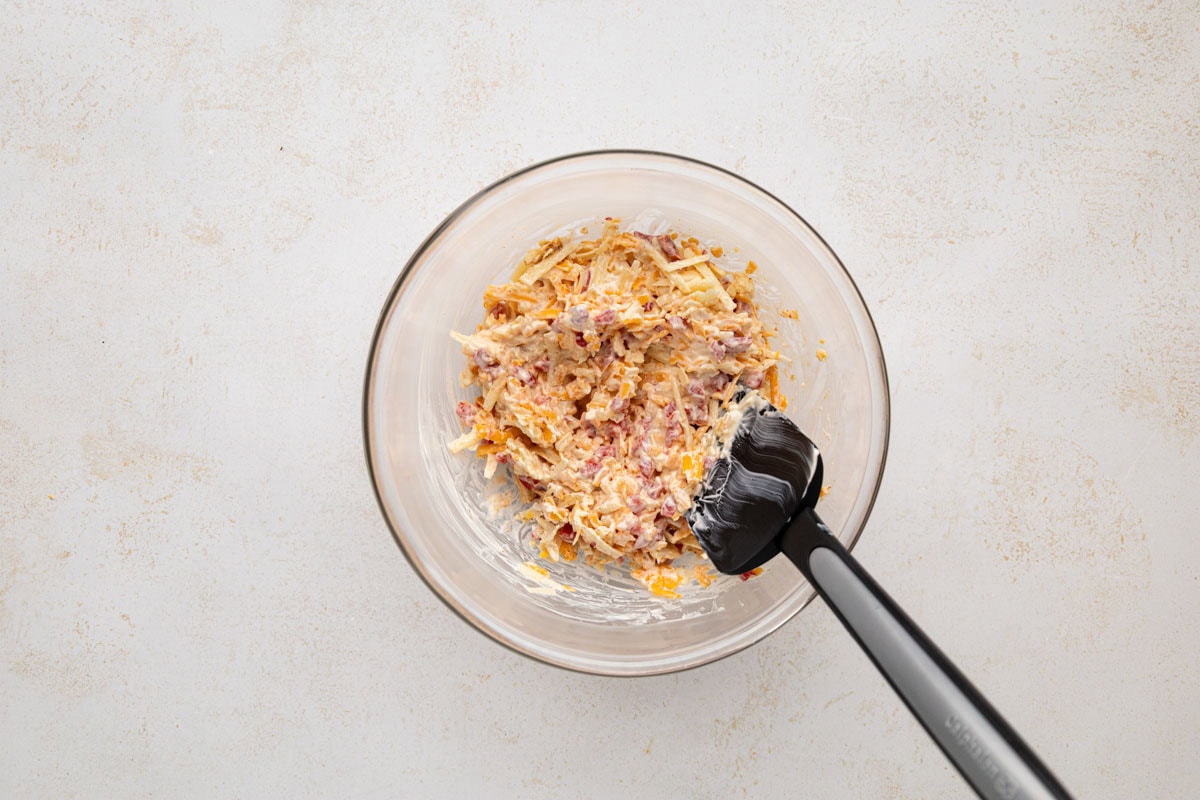 Ingredients for pimento cheese stirred together with a spatula in a glass bowl.