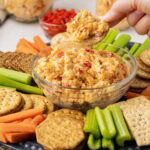 A hand picking up a cracker with pimento cheese spread on top from a tray with crackers and veggies.