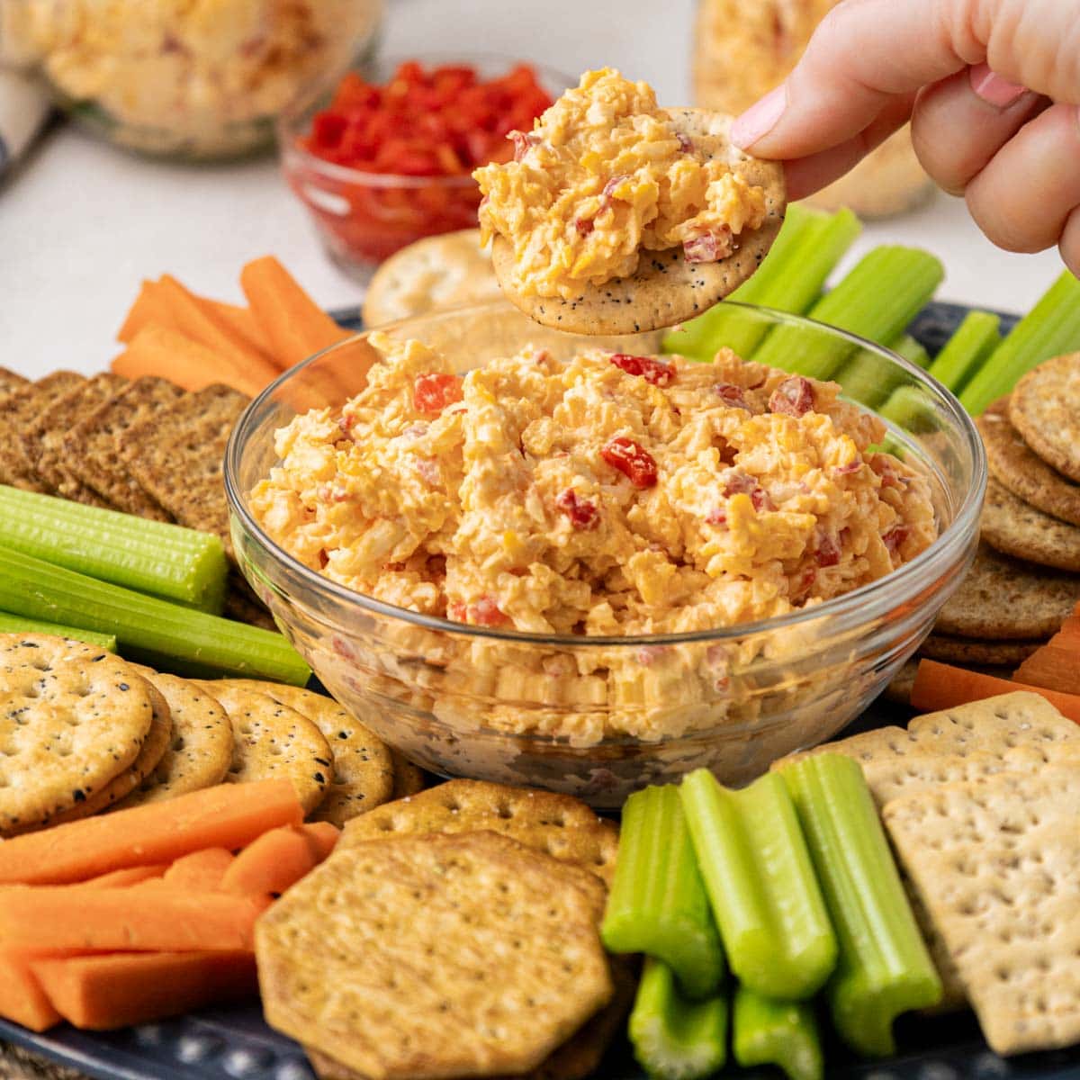 A hand picking up a cracker with pimento cheese spread on top from a tray with crackers and veggies.