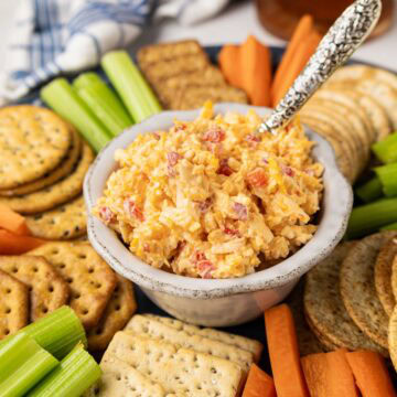 Pimento cheese in a bowl on a round wooden tray with crackers, celery and carrots.