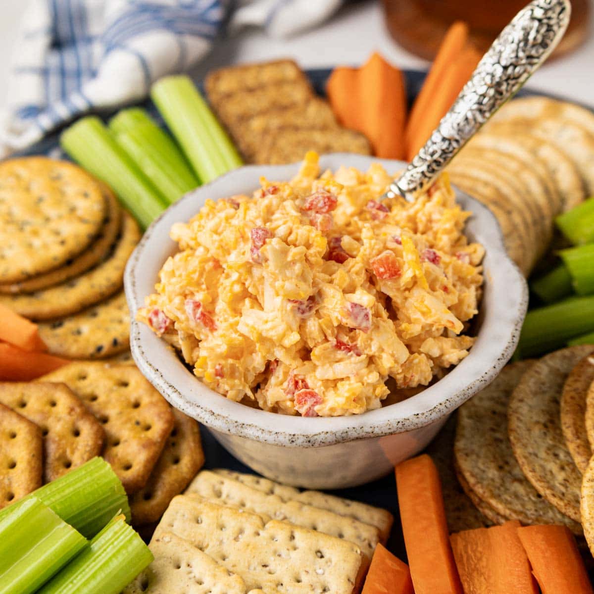 Pimento cheese in a bowl on a round wooden tray with crackers, celery and carrots.