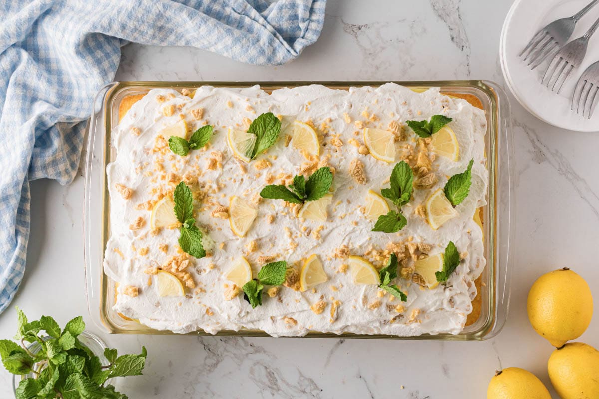 Lemon pudding poke cake topped with whipped cream icing and garnished with lemon wedges, crushed cookies, and mint leaves.