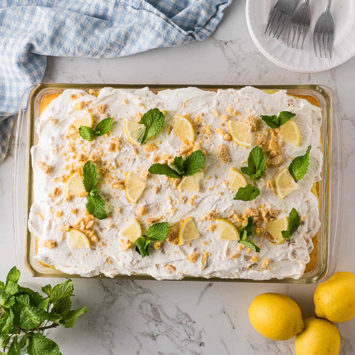 Lemon pudding poke cake topped with whipped cream icing and garnished with lemon wedges, crushed cookies, and mint leaves.
