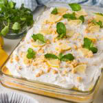Lemon pudding poke cake topped with whipped cream icing and garnished with lemon wedges, crushed cookies, and mint leaves.