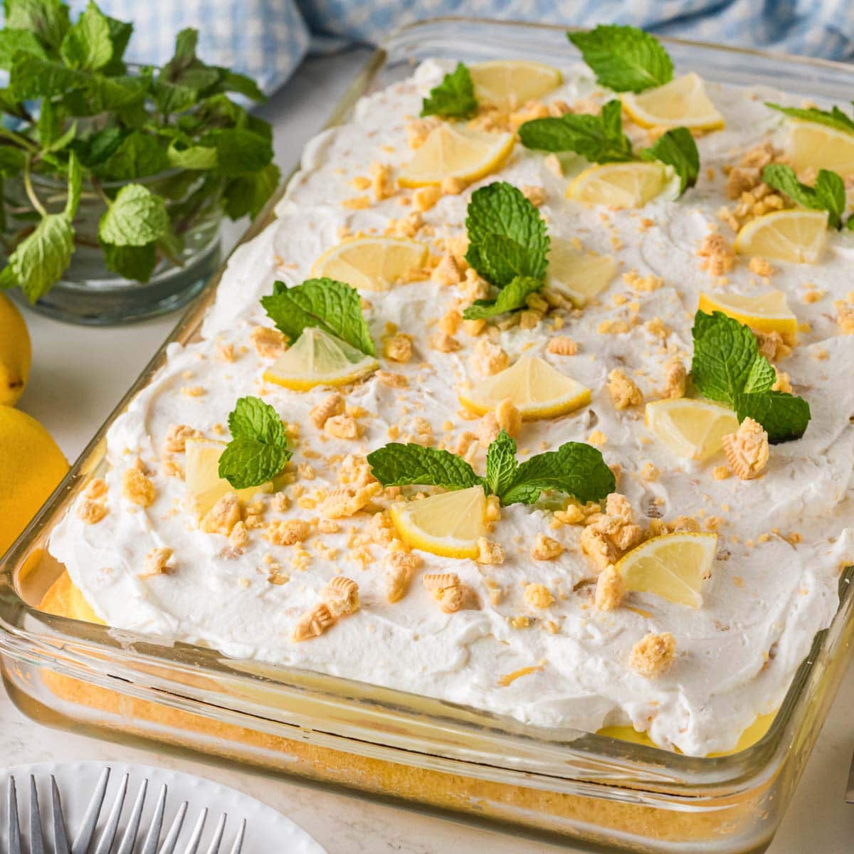 Lemon pudding poke cake topped with whipped cream icing and garnished with lemon wedges, crushed cookies, and mint leaves.