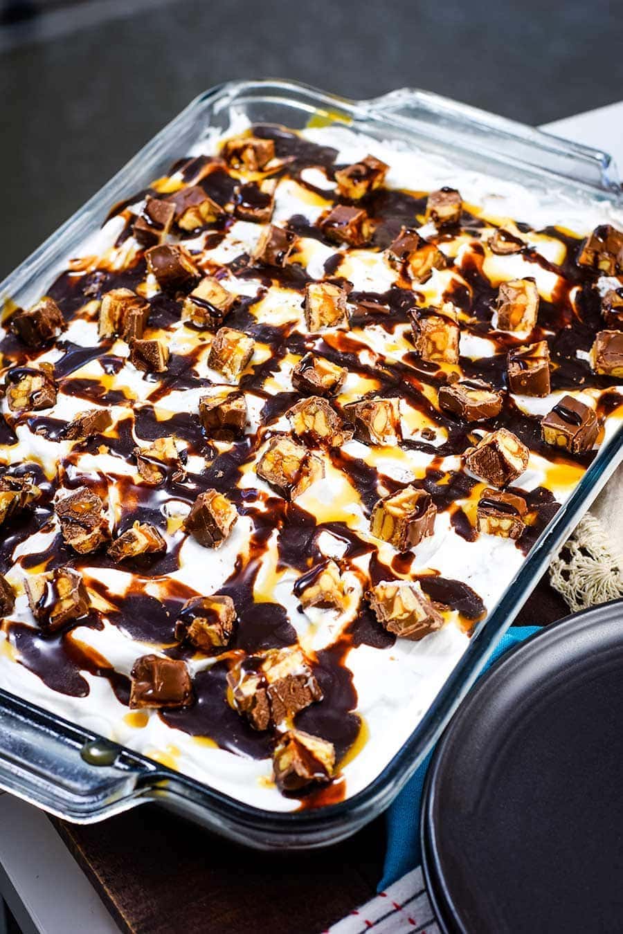 Close up of Snickers Poke Cake in a casserole dish topped with chopped Snicker bars, chocolate sauce, and caramel sauce