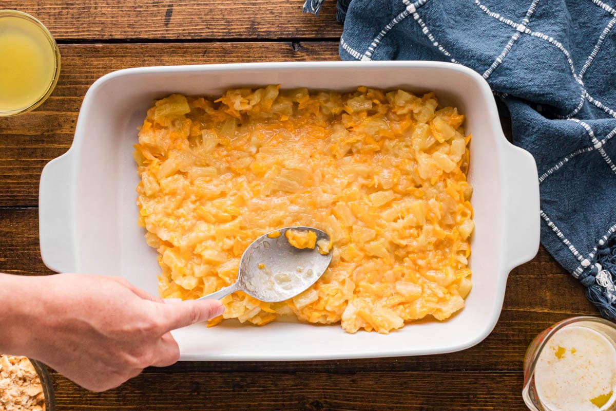 A spoon smoothing the pineapple cheese mixture into the bottom on a casserole dish.