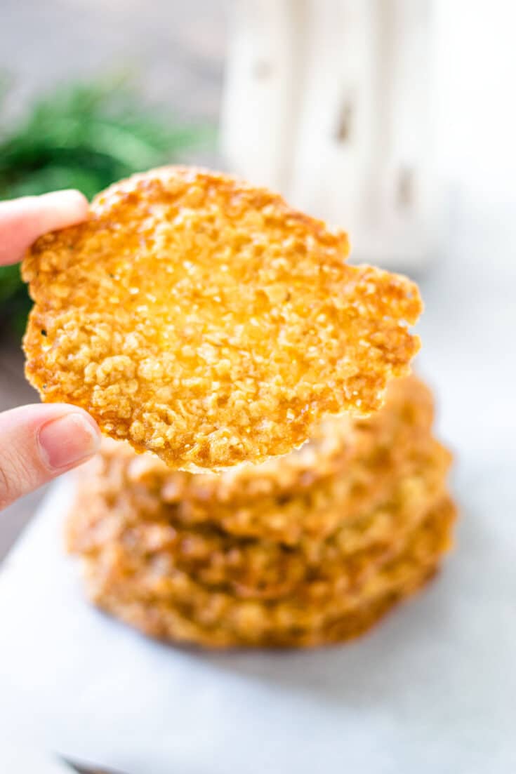 a hand holding up one Oatmeal Lace Cookie with a stack of these Easy Oatmeal Lace Cookies in the background