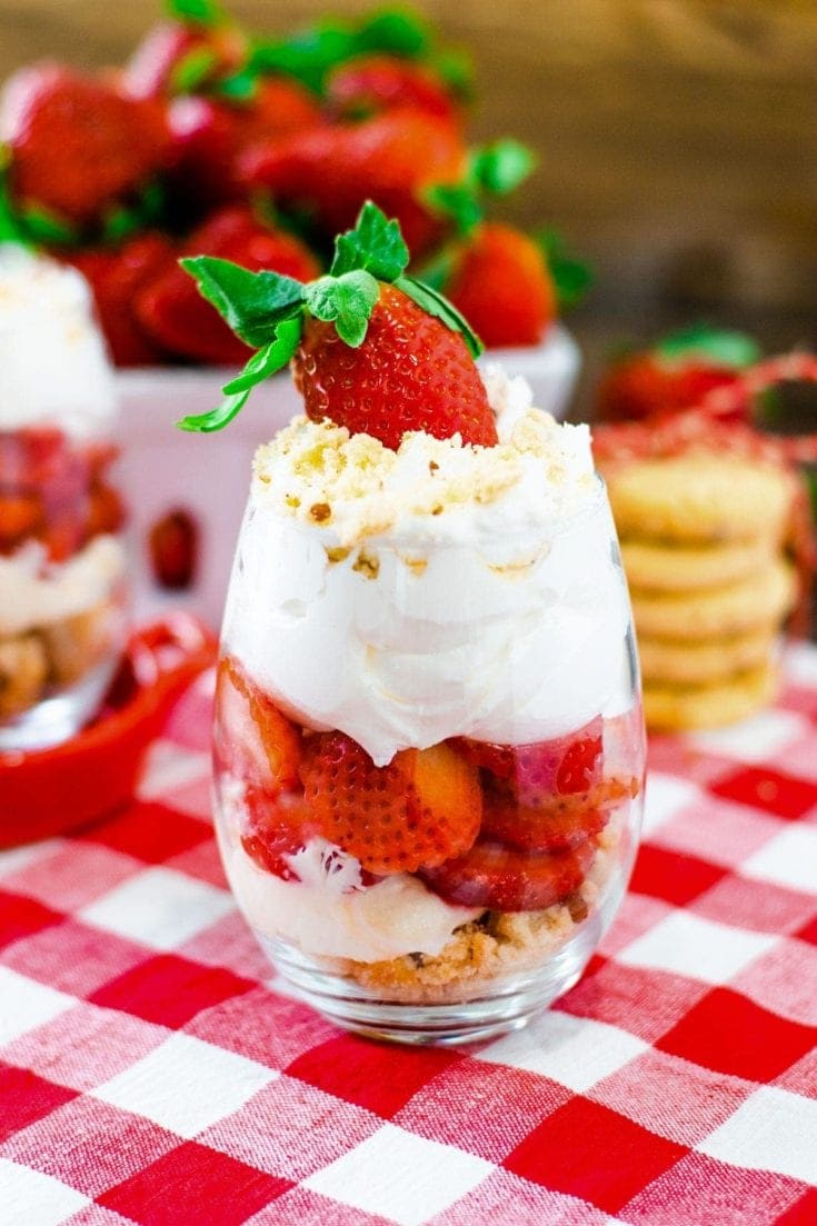 Strawberry Parfait with layers of shortbread cookie crumbs, sliced strawberries and whipped cream on a red and white checkered table cloth.