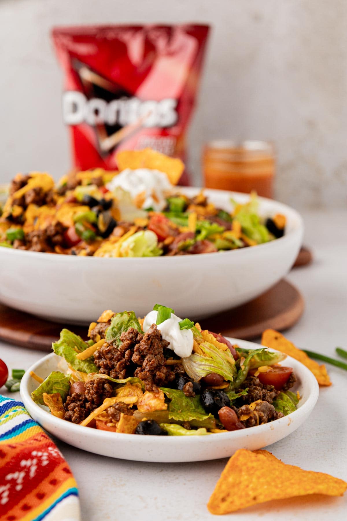 A Dorito taco salad on a white serving plate with the bowl of salad in the background and a bag of Doritos.