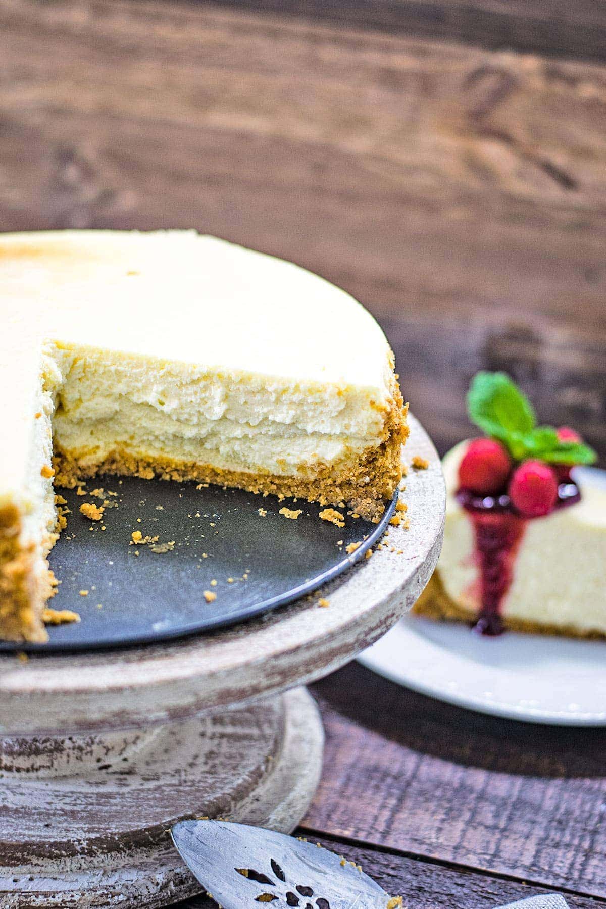 A cut cheesecake on a cake pedestal with a slice of cheese topped with raspberry sauce in the background.