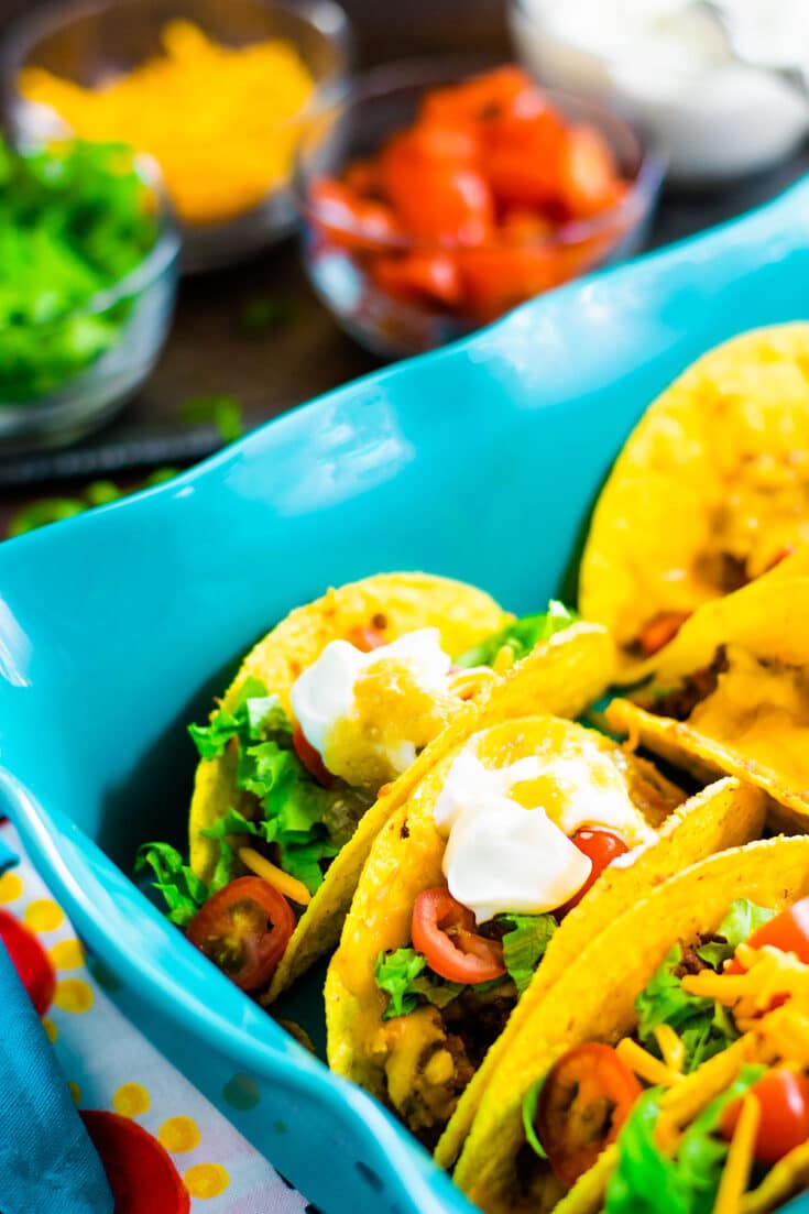 A blue baking dish with baked tacos garnished with lettuce, tomatoes, and sour cream.