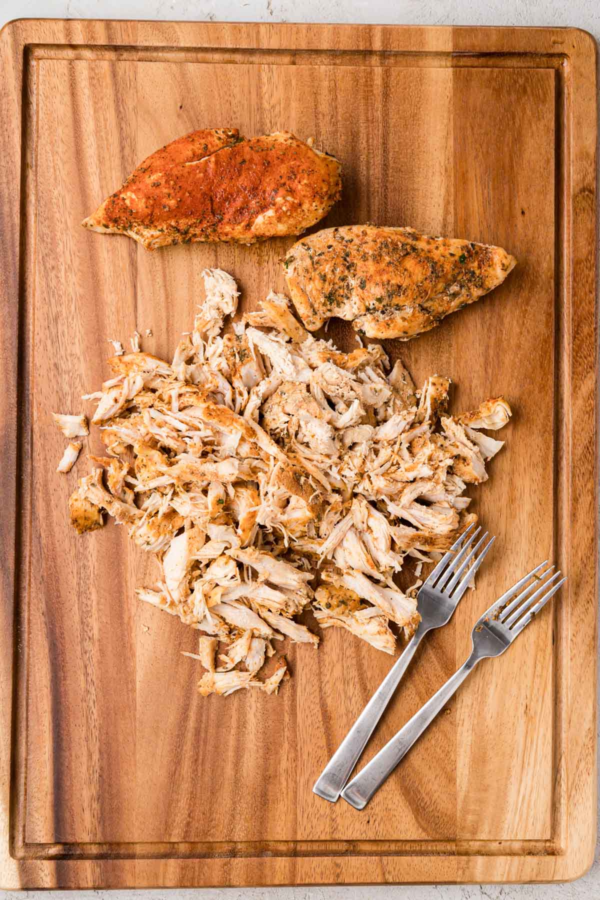 A wooden board with 2 chicken breast and 2 forks along with shredded chicken.