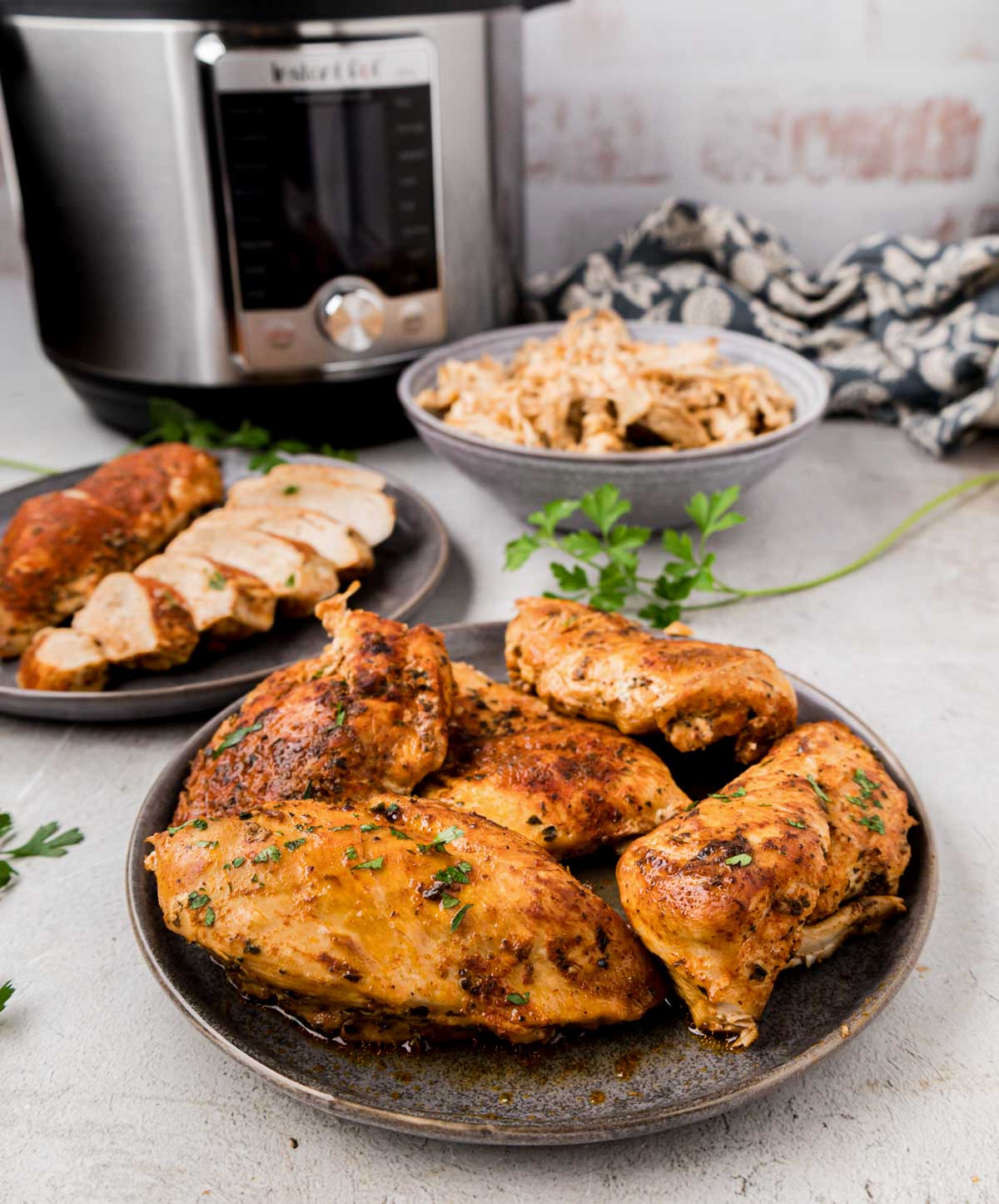 A plate of whole chicken, one with sliced chicken and a bowl of shredded chicken with the instant pot it was cooked in in the background.