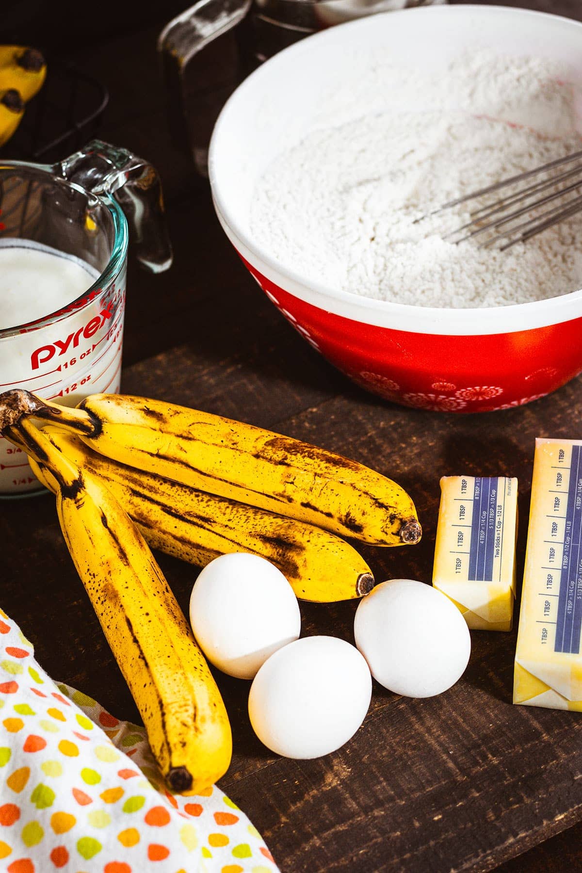 Ingredients for Banana Cake. Ripe bananas, eggs, butter, flour mixture in a red bowl and a measuring cup of buttermilk.