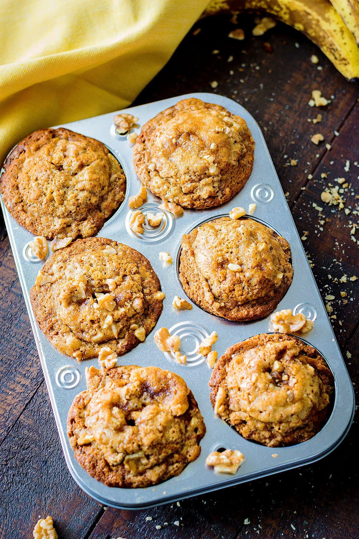 Muffins in a muffin tin surrounded by scatter walnuts on a wooden table with yellow napkin and bananas in the background.