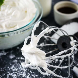 A whisk with whipped cream laying on black table scatter with sugar and a bowl of whipped cream in the background.
