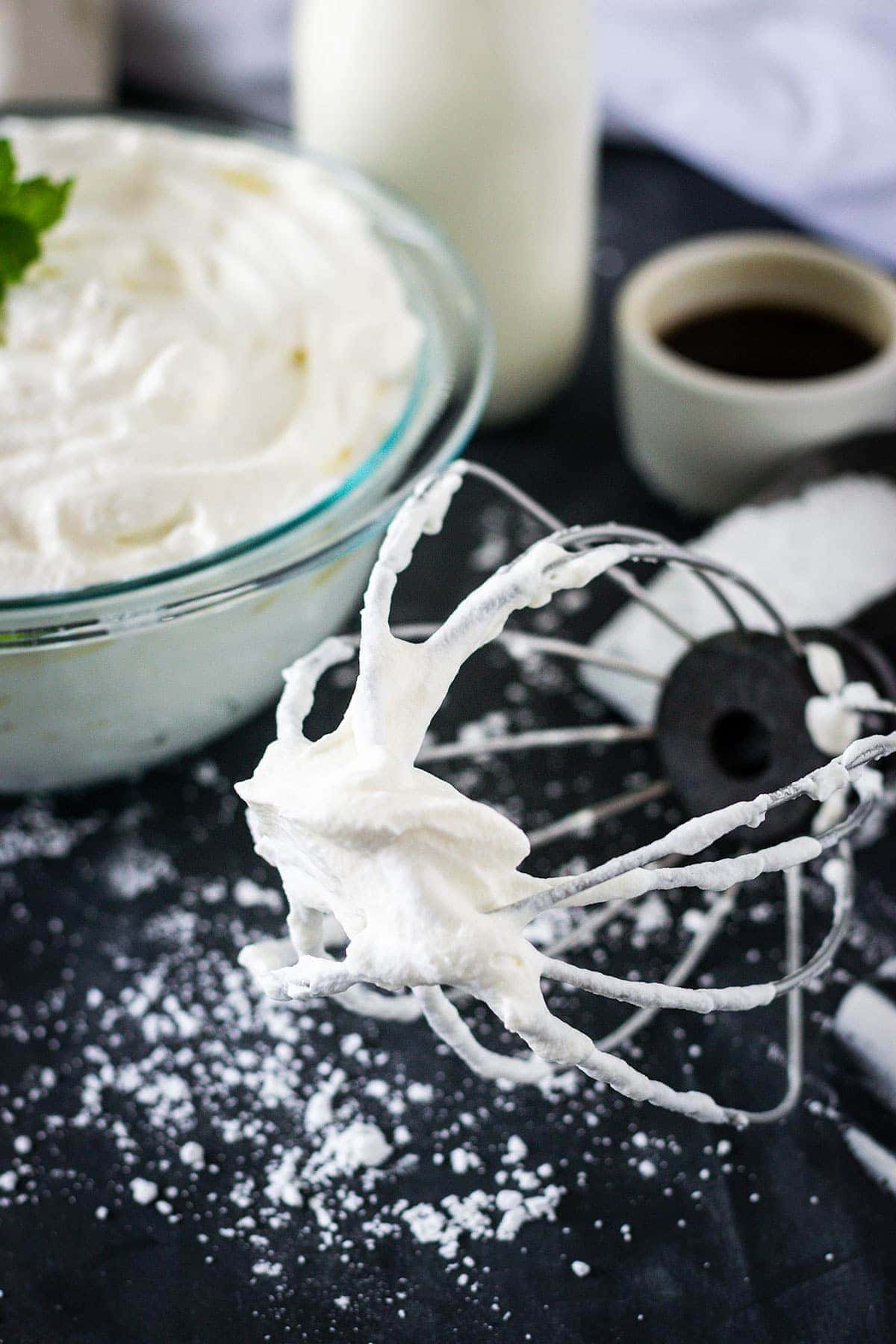 A whisk with whipped cream laying on black table scatter with sugar and a bowl of whipped cream in the background.