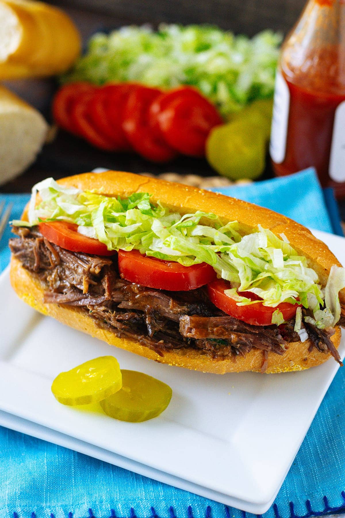 A roast beef debris sandwich sitting on top of a white plate served with pickles. 