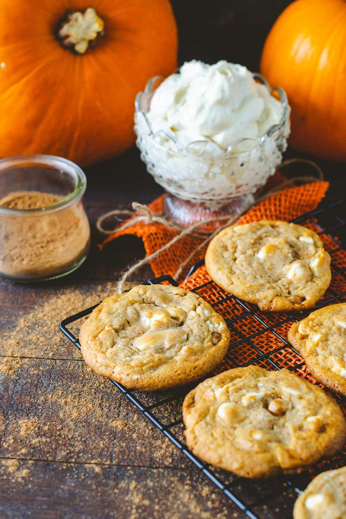 White Chocolate Caramel Pumpkin Spice Cookies on a cooling rack with White Chocolate Buttercream icing in a glass dish and orange pumpkins  in the background. 