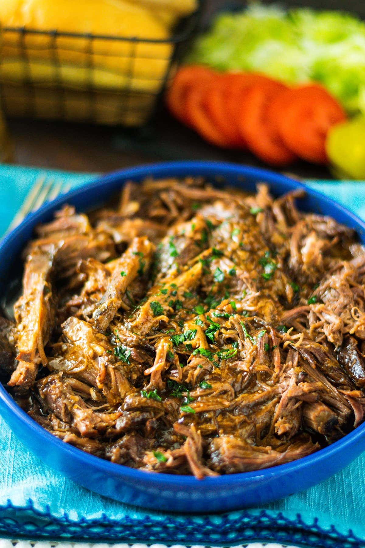 A blue bowl filled with shredded roast beef debris with tomatoes in the background.