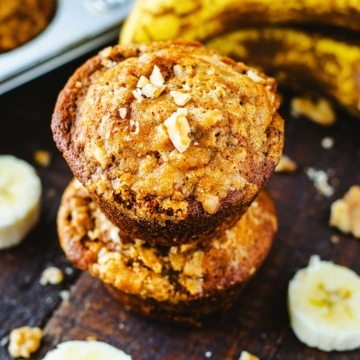 Two stacked Banana Nut Muffins on a wooden serving tray with ripened bananas and a tray of muffins in background.