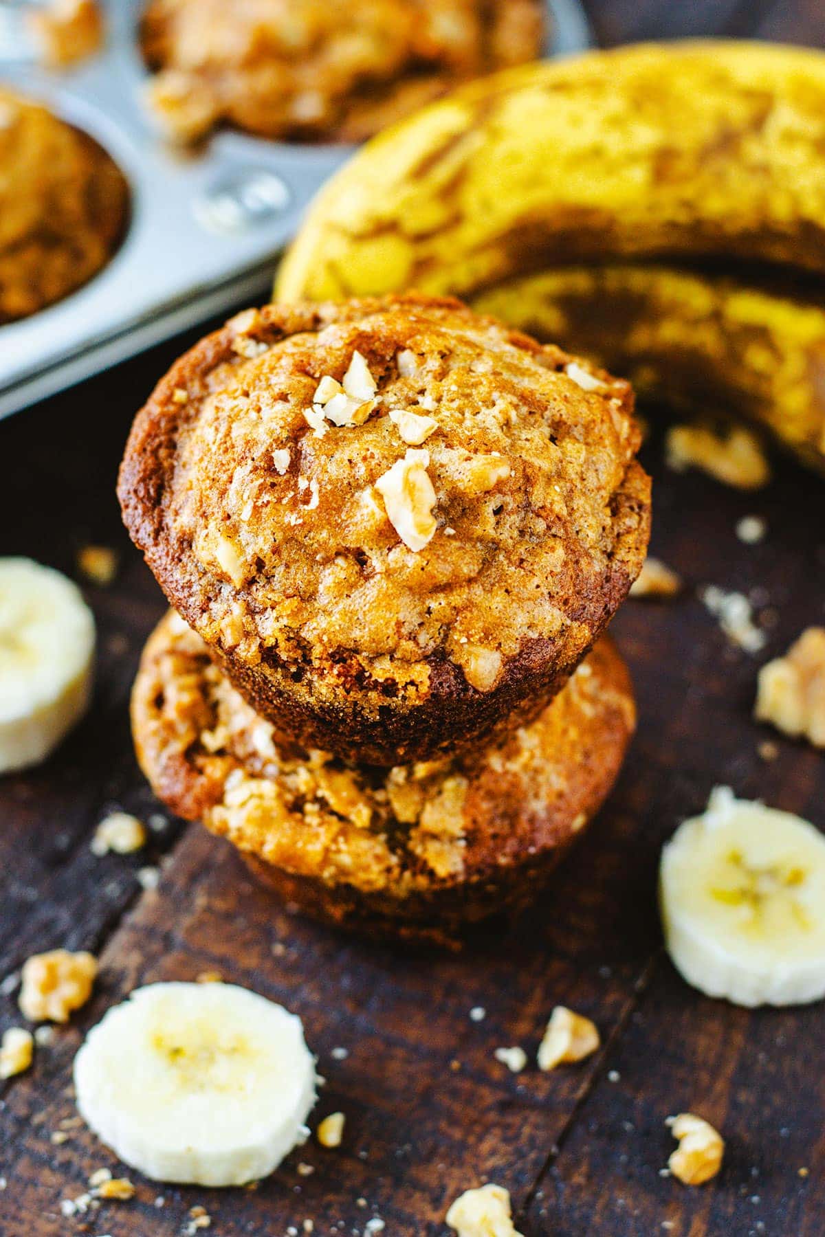 Two stacked Banana Nut Muffins on a wooden serving tray with ripened bananas and a tray of muffins in background.