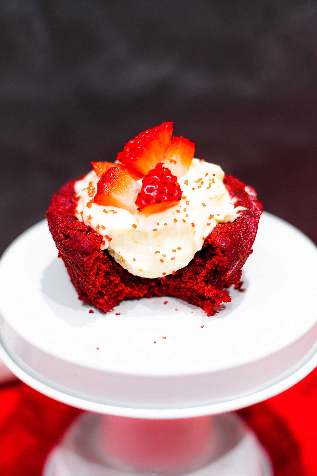 Red Velvet Brownie Cup on a white mini cake stand. Filled with no bake cheesecake and topped off with chopped strawberries and red sugar 
sprinkles.