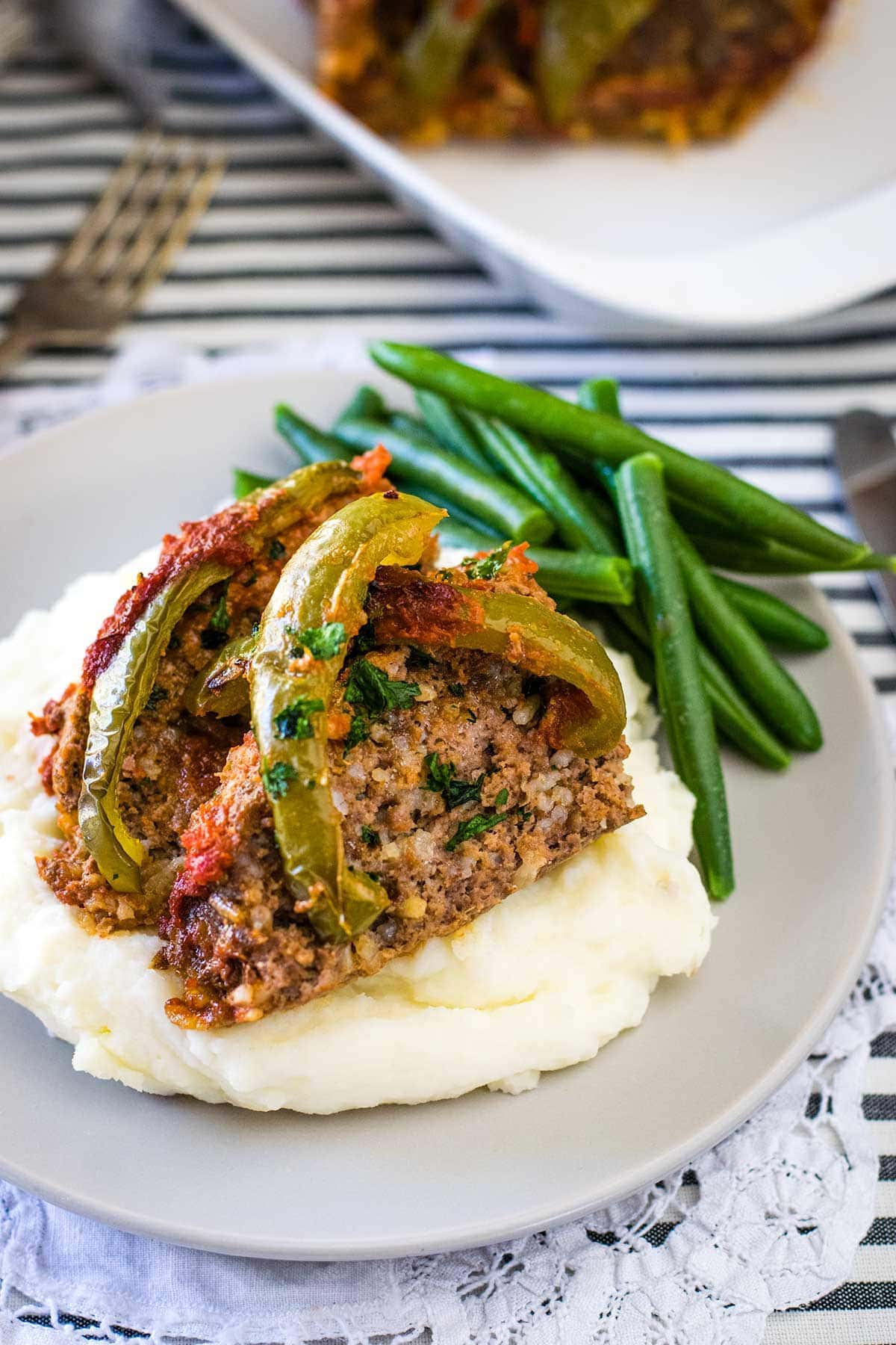 Meatloaf topped with peppers and tomatoes on a bed of mashed potatoes and a side of green beans.