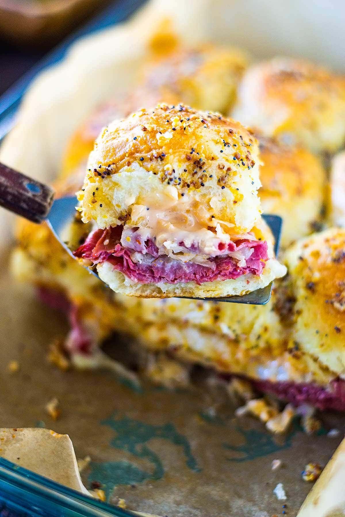 A tray of hawaiian roll reuben sandwich sliders with one removed on a serving spatula for serving.