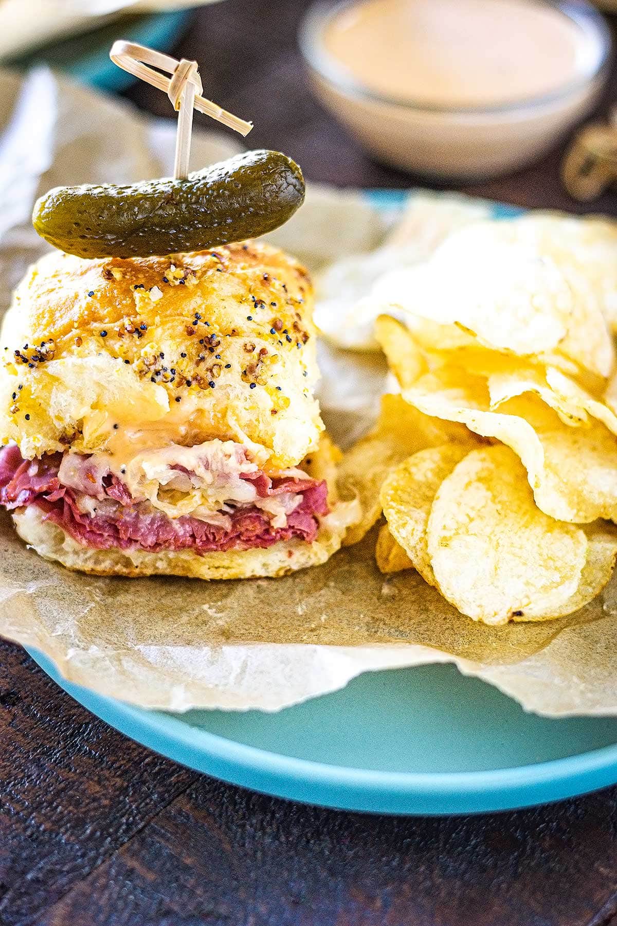 A baked reuben sandwich slider topped with a pickle along with kettle chips on a plate lined with parchment paper.