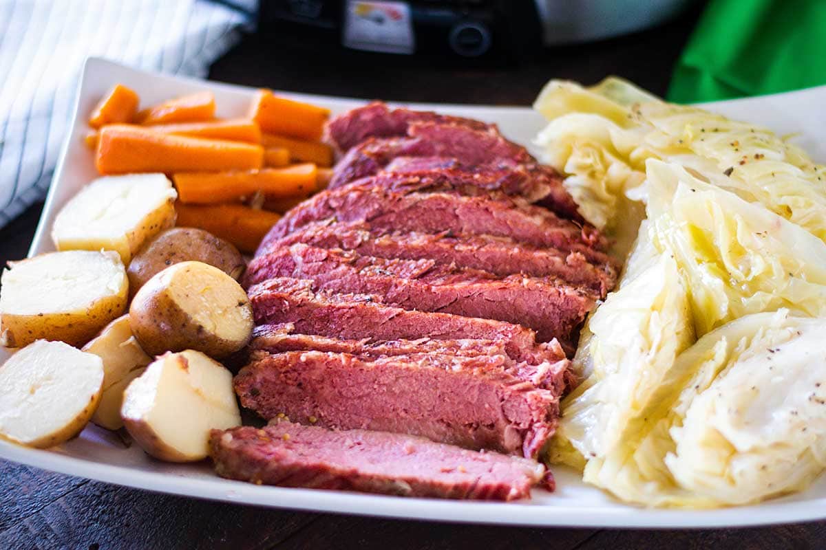 Sliced corned beef on a serving dish with potatoes, cabbage and carrots with slow cooker with the background.