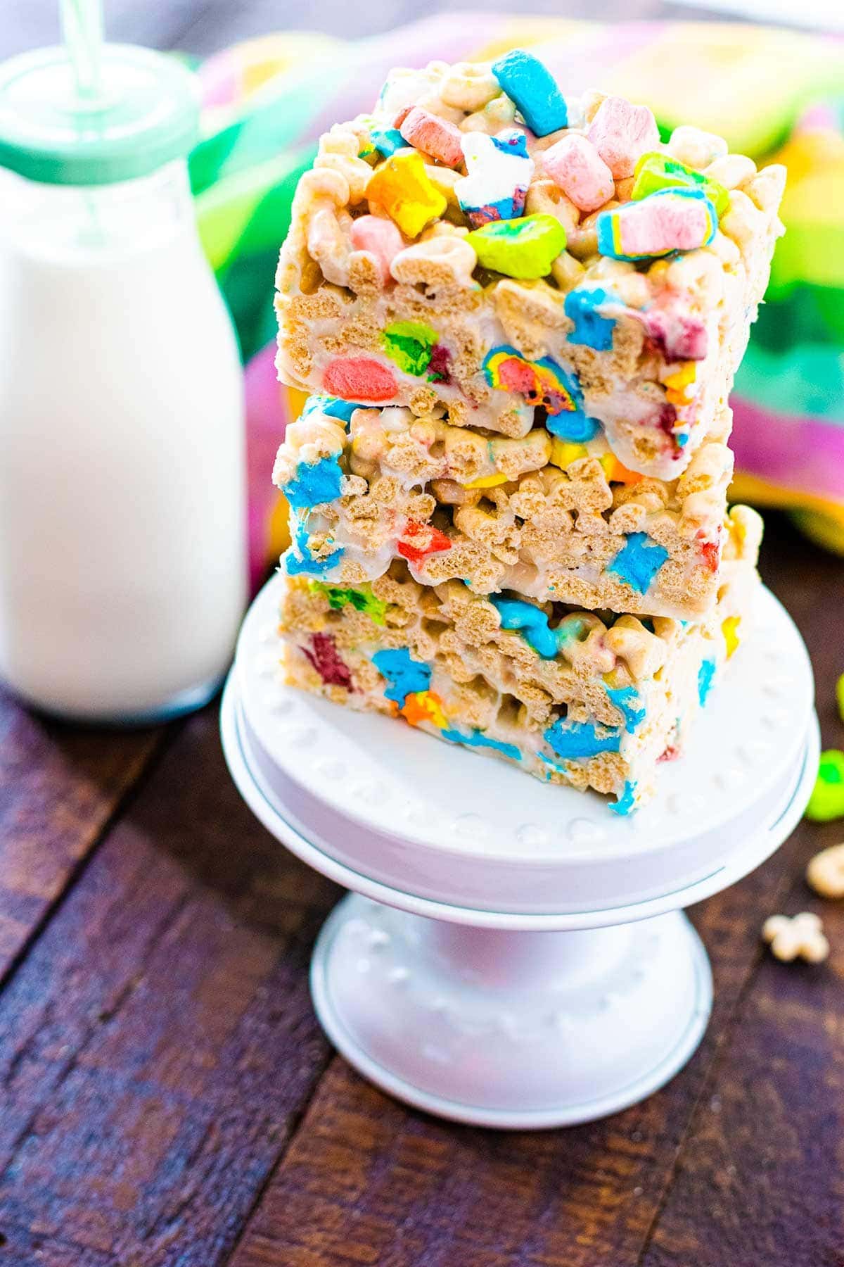 Lucky Charms Treat cut and stacked on a mini cake stand with milk and a vibrant rainbow hand towel in the background. 